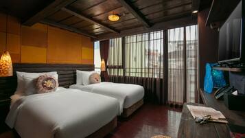 A hotel room with two beds, a wooden ceiling, a large window with curtains, a desk with a lamp, and a TV.