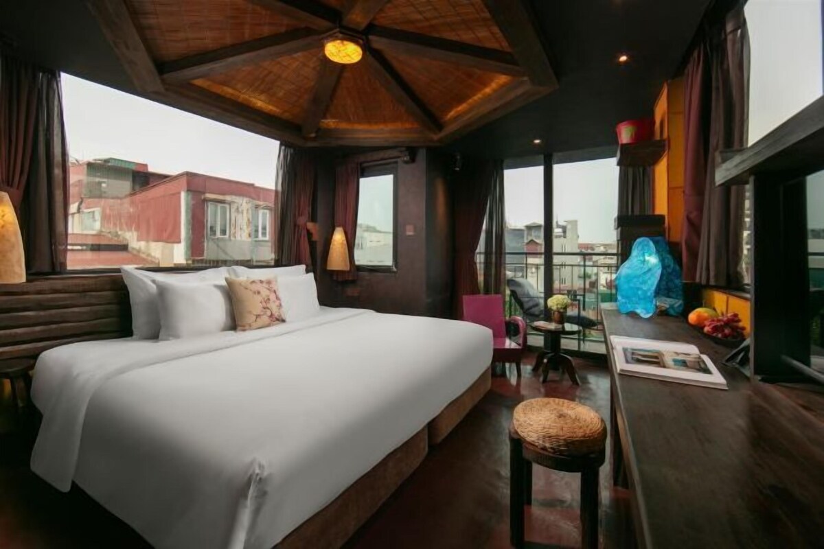 Habitación de hotel con una cama grande, una mesita con una silla y vistas a edificios a través de las ventanas.