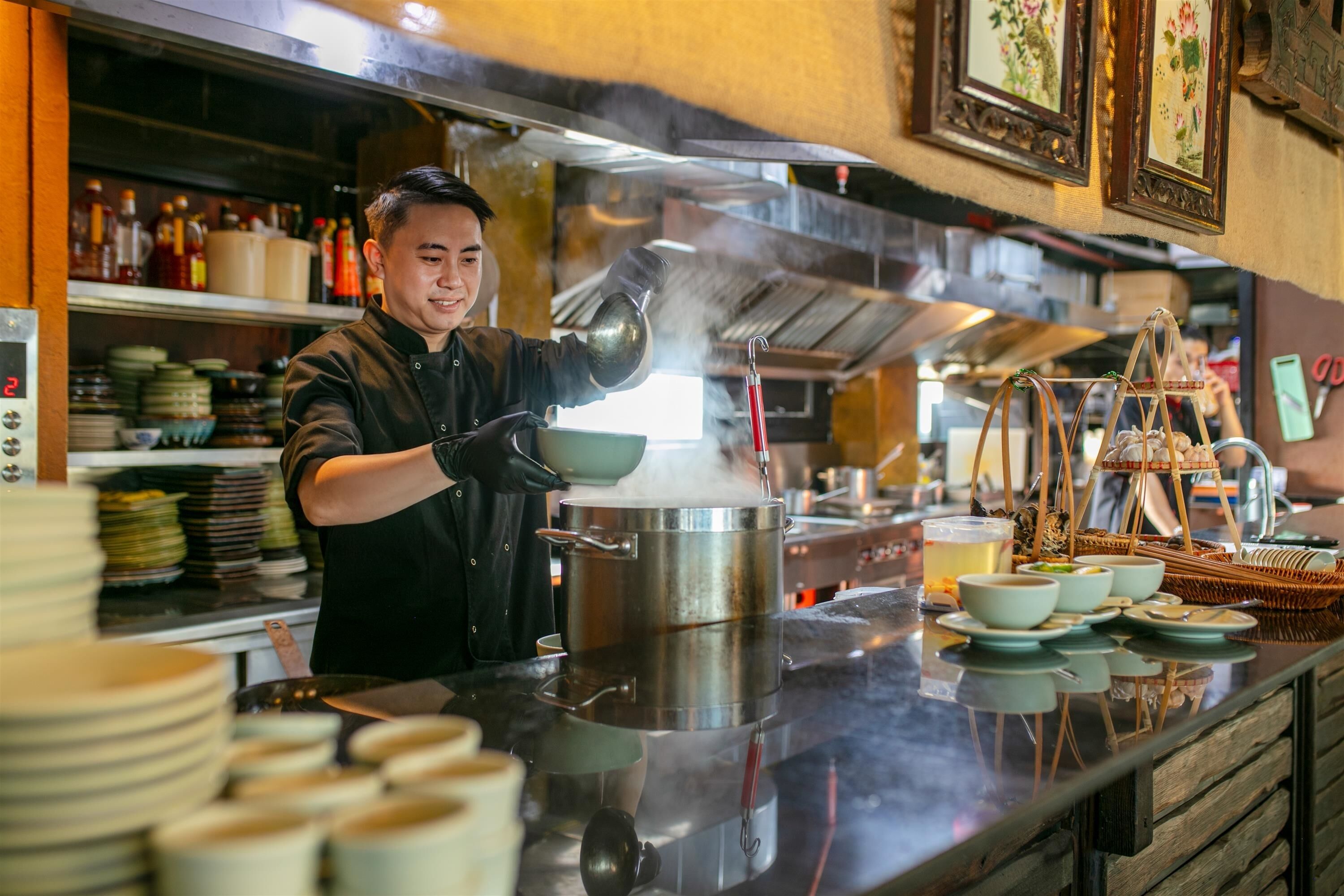 A chef pouring a hot liquid from a pot into a steaming container on a kitchen counter.