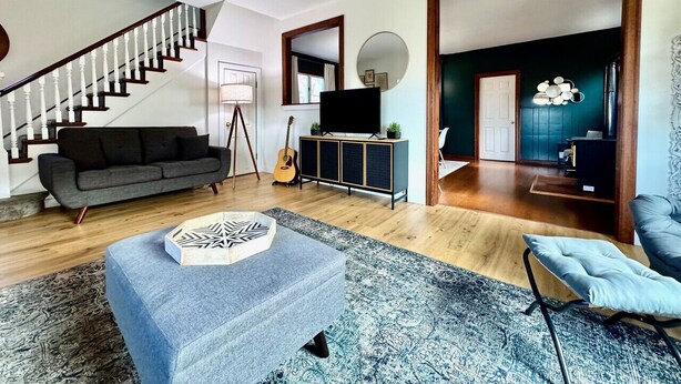 Una sala moderna con escalera, un sofá gris, una mesa de centro, un mueble para televisor y un comedor visible a través de una puerta abierta.
