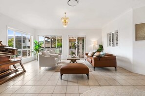 A spacious living room with a piano, two sofas, a coffee table, and a chandelier. - Peaceful Family Home: 4-Bed, Pool, Alfresco Dining (Bridgeman Downs)