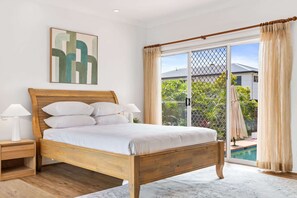 A bedroom with a wooden bed, two bedside tables, a large window with a view of a swimming pool and greenery, and a ceiling light. - Peaceful Family Home: 4-Bed, Pool, Alfresco Dining (Bridgeman Downs)