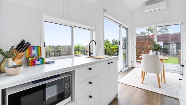 A modern kitchen with a large window, a built-in microwave, and a dining area with a white table and chairs. - Cute 1-Bed Tiny House with Garden in Nature (Bridgeman Downs)
