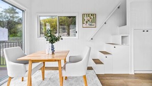 A modern kitchen with white cabinetry, a wooden dining table, and a view of the outdoors through large windows. - Cute 1-Bed Tiny House with Garden in Nature (Bridgeman Downs)