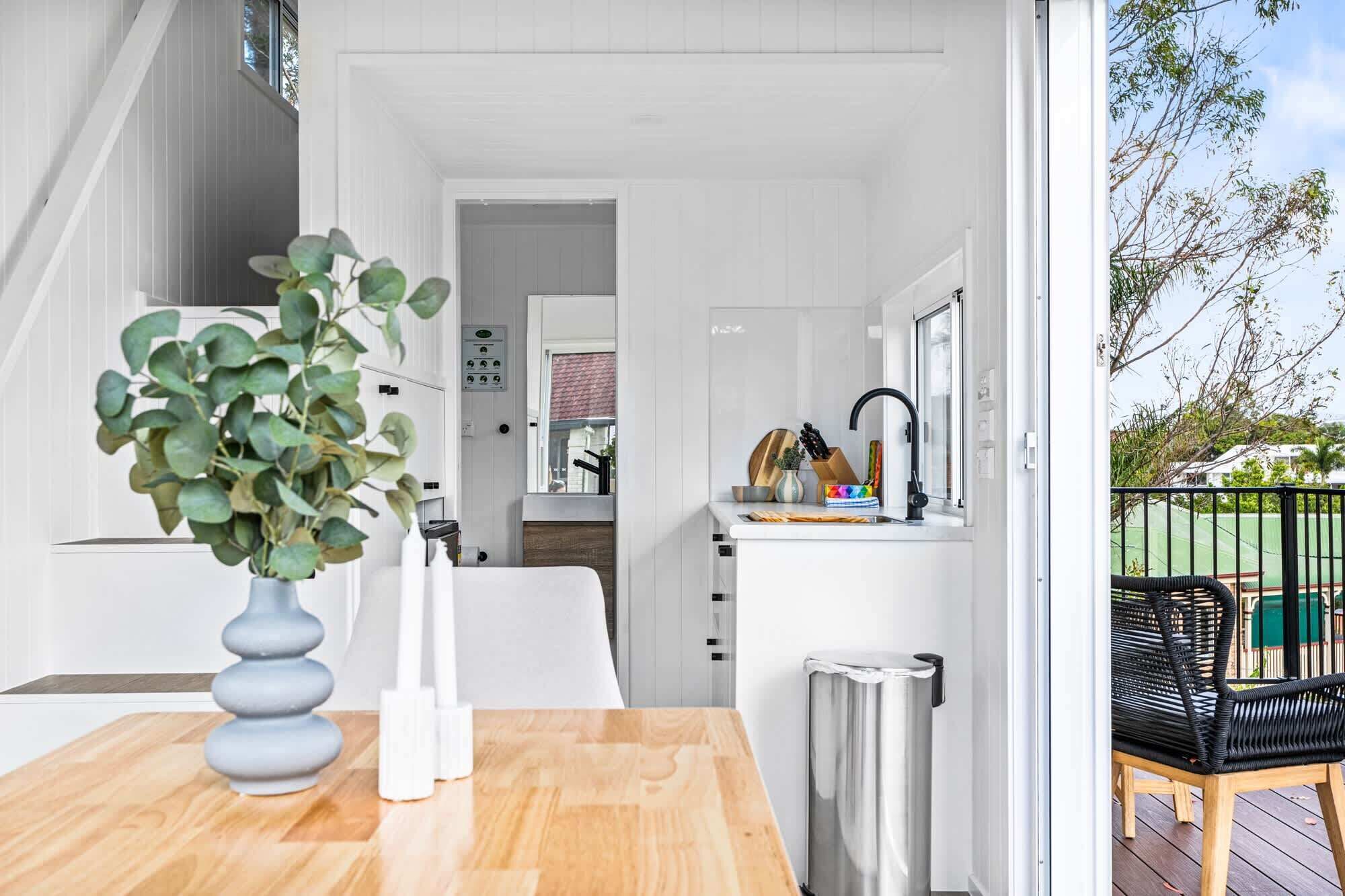 Une cuisine moderne avec des meubles blancs, un évier en acier inoxydable et un petit îlot central. On aperçoit une plante en pot sur la gauche et, à travers la fenêtre, une vue sur l’extérieur.