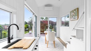 A modern kitchen with a large window, a sink with a wooden cutting board, and a dining area with a white table and chairs. - Cute 1-Bed Tiny House with Garden in Nature (Bridgeman Downs)