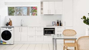 A modern kitchen with white cabinets, a built-in washing machine, a glass dining table, and a wicker chair. - Chic 1-Bed Escape with Parking Bridgeman Downs (Bridgeman Downs)
