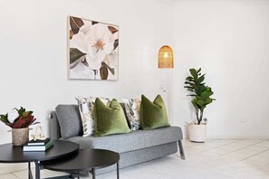 A living room with a grey sofa, green cushions, a round table, a potted plant, and a wall-mounted light fixture.