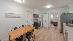 Une cuisine compacte avec des meubles blancs, un four à micro-ondes, un four et un réfrigérateur. On aperçoit une table à manger avec des chaises au premier plan.