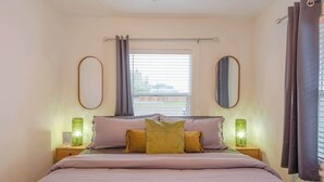 Une chambre à coucher comprenant un lit, deux tables de chevet, une fenêtre avec des rideaux et deux miroirs fixés au mur.