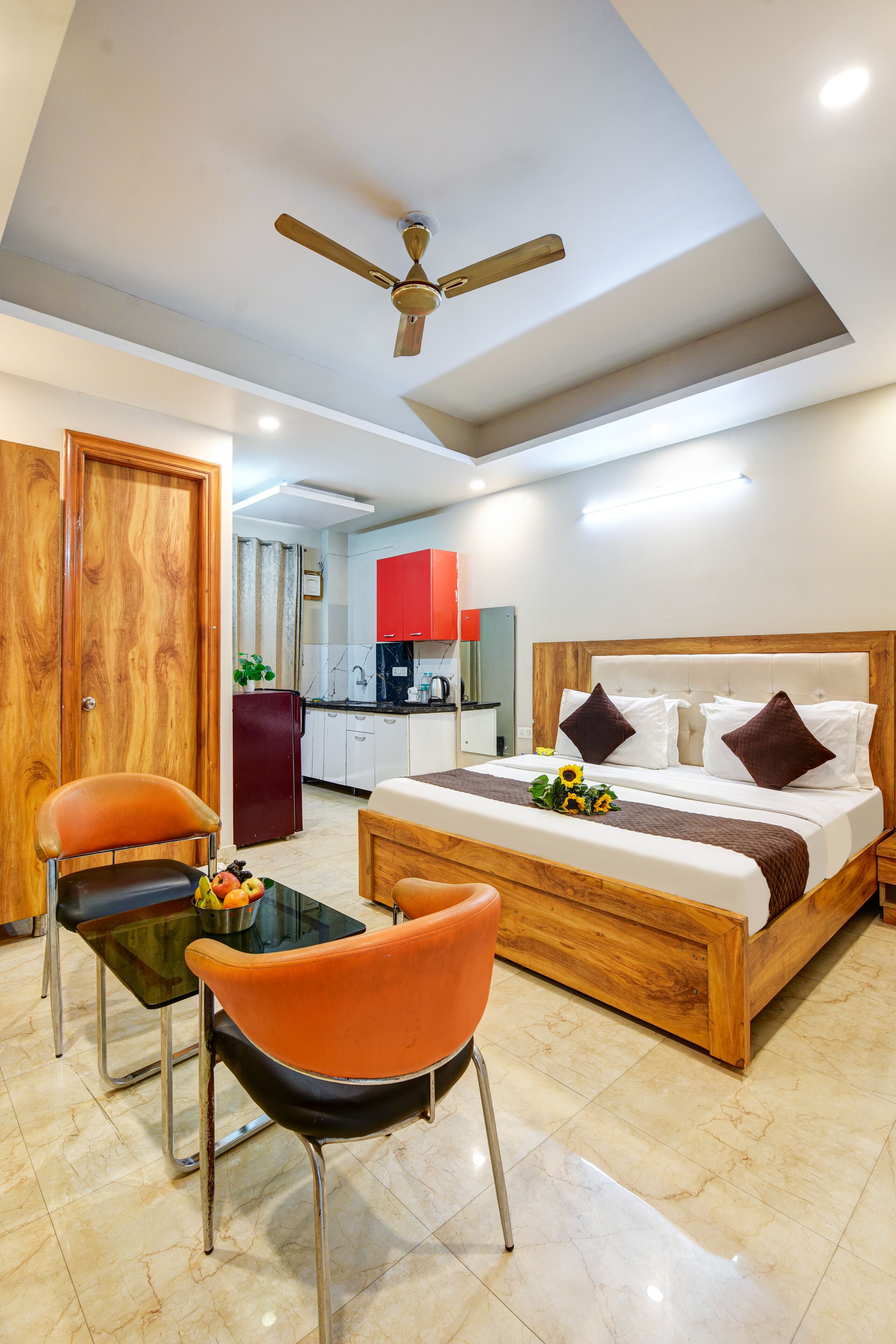 A modern hotel room with a wooden bed, orange chairs, a small table, a ceiling fan, and a kitchen area in the background.