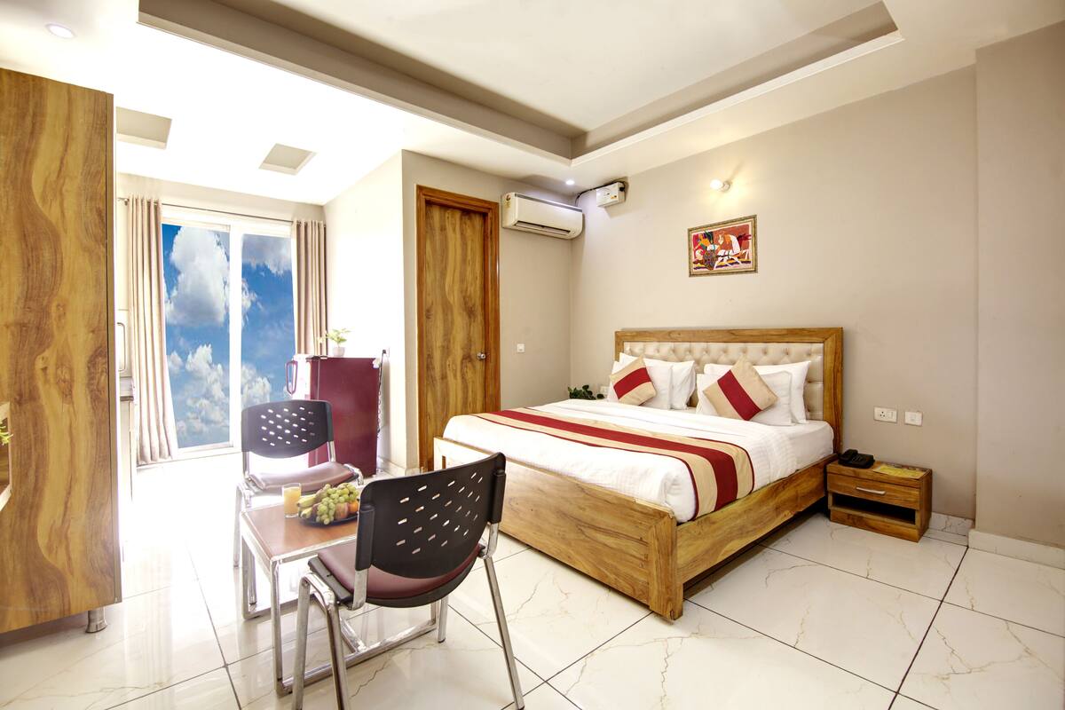 A bedroom with a wooden bed, a desk with a chair, a small bedside table, and a view of the sky through a window.