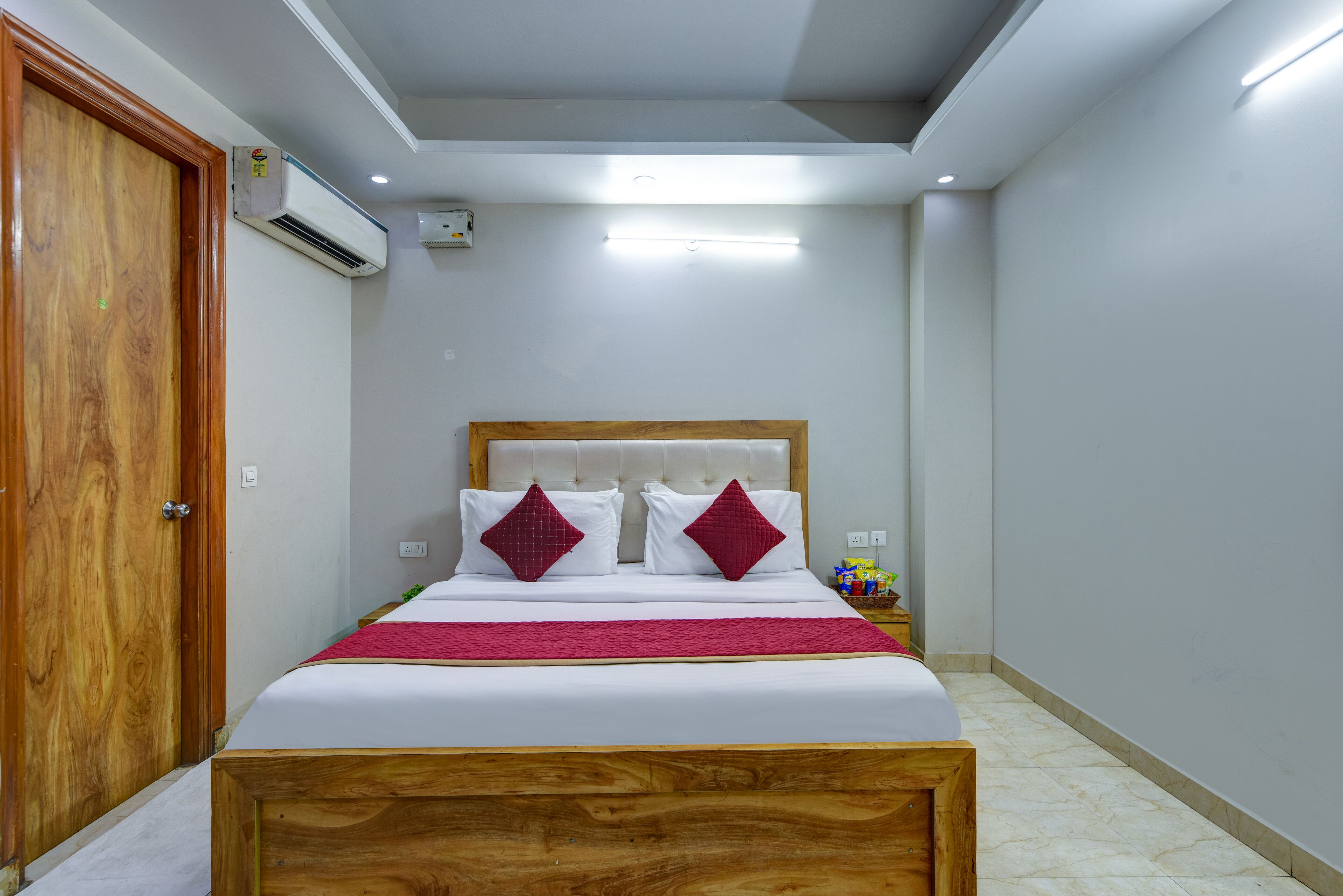 Un dormitorio ordenado con estructura de cama de madera, cubrecama blanco y dos almohadas rojas.