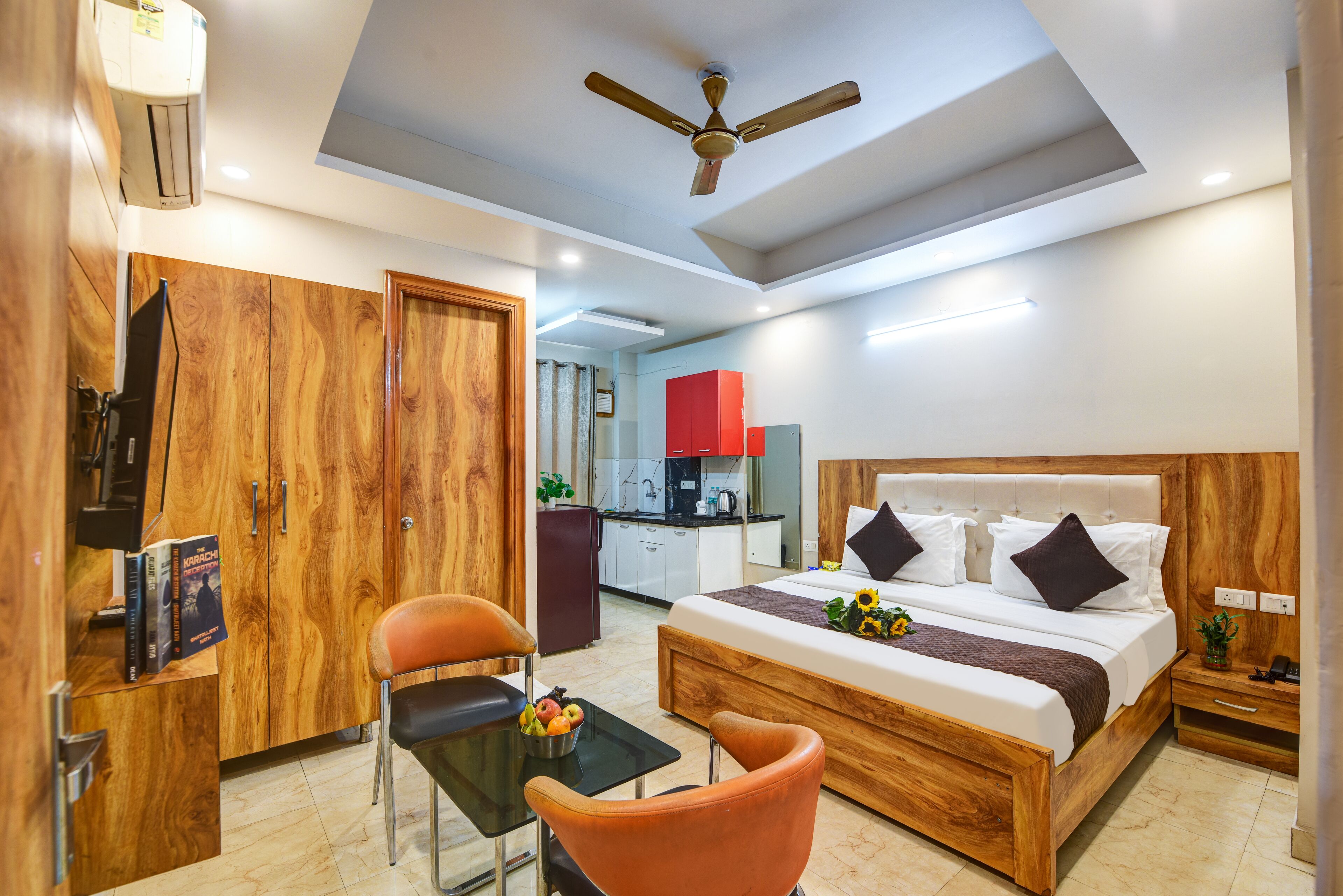 Une chambre d’hôtel moderne avec un lit en bois, un bureau avec une chaise, un ventilateur de plafond et, au fond, une kitchenette.