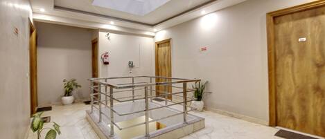 Un hall de hotel con barandilla de vidrio, plantas en macetas, suelo de mármol y puertas de madera.