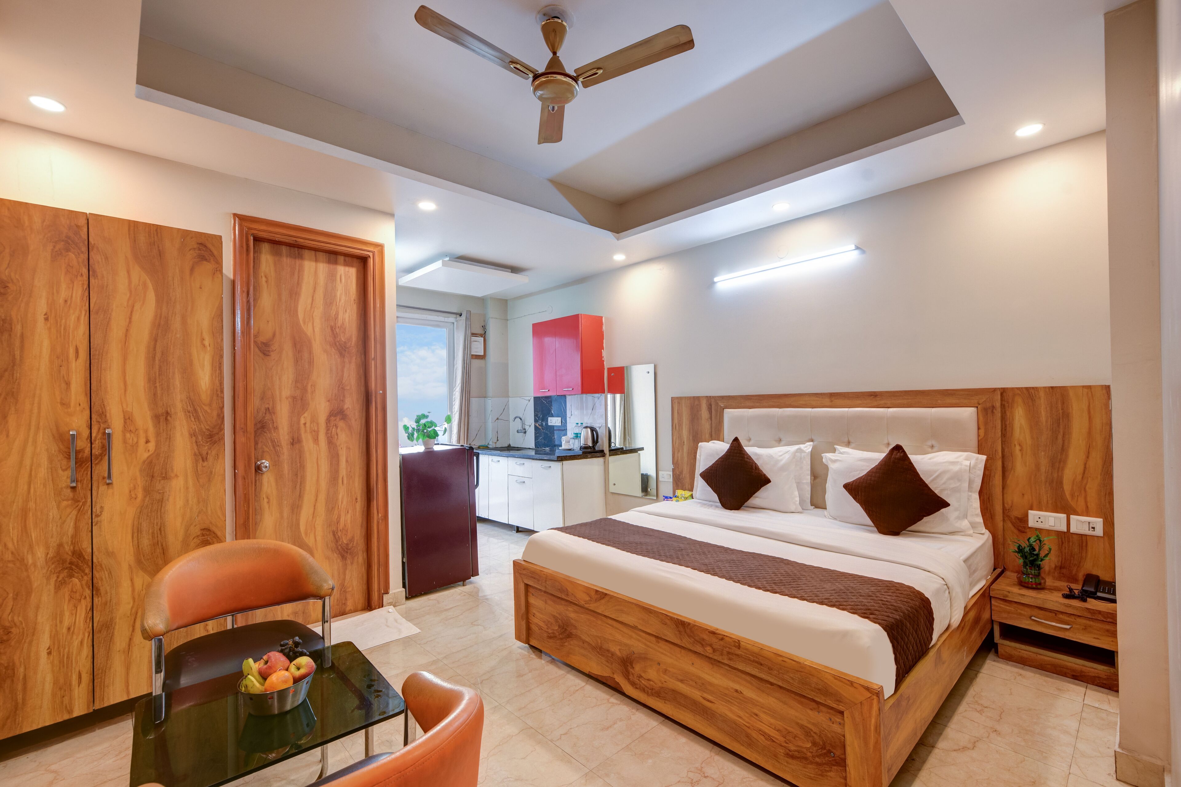 A modern hotel room with a wooden bed, a glass table with fruits, a wooden wardrobe, and a kitchenette in the background.