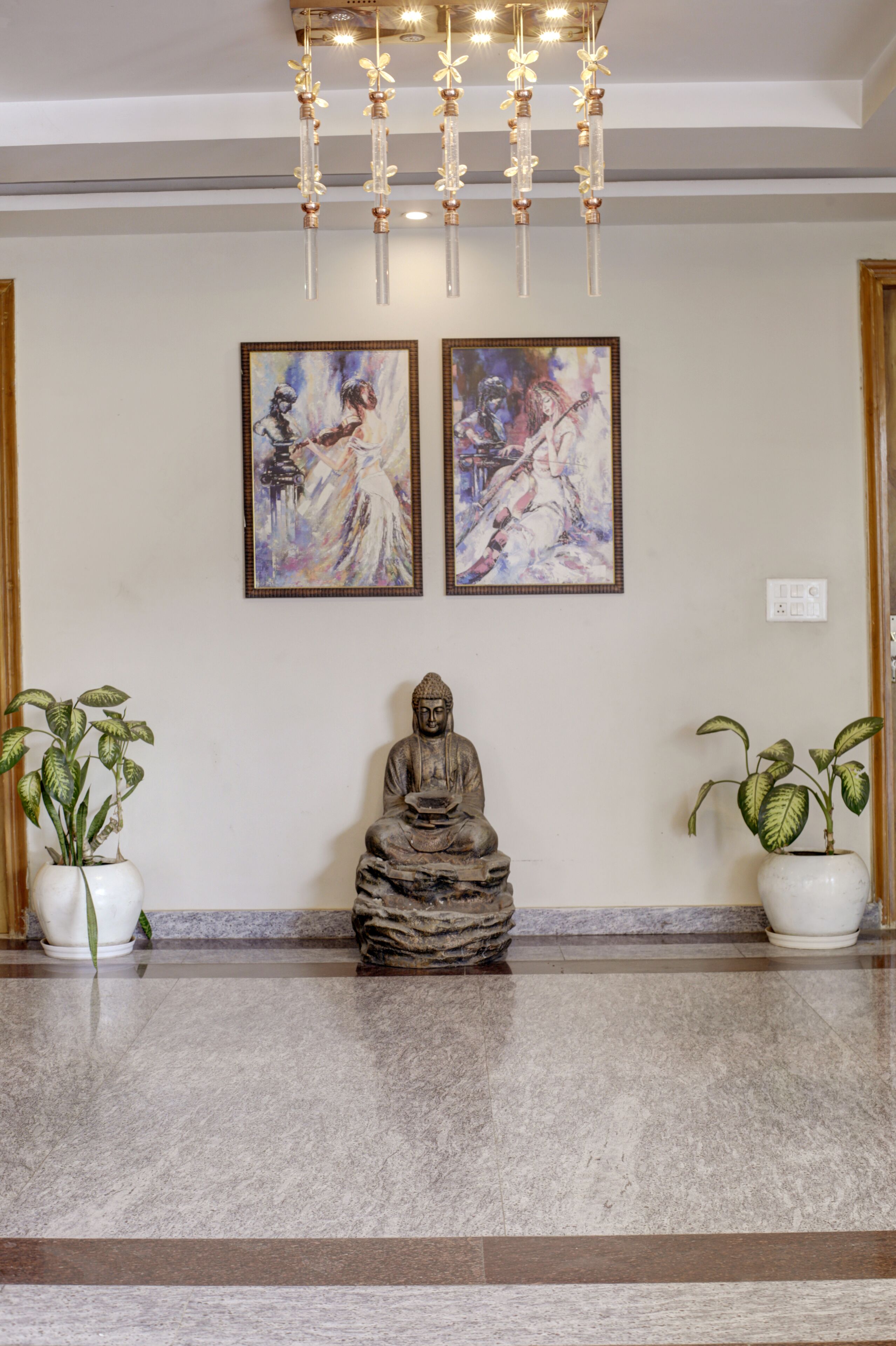 A statue of Buddha in a meditative pose, flanked by two potted plants, with two framed paintings above.
