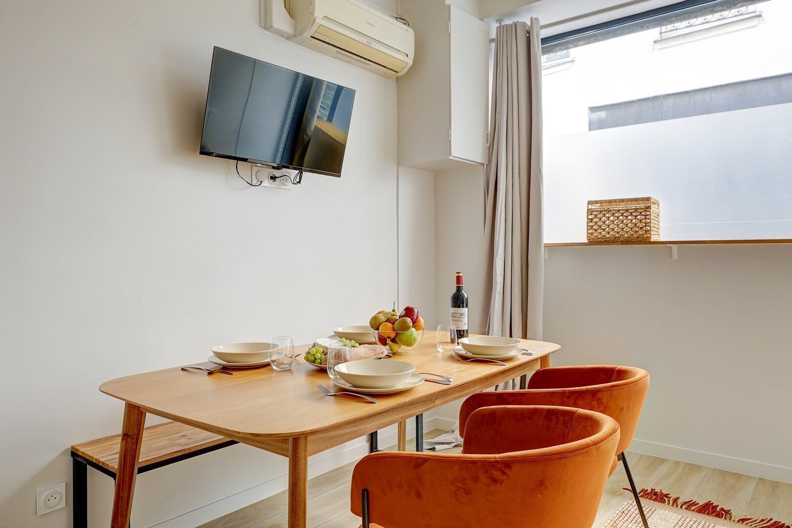 A dining area with a wooden table, orange chairs, a TV mounted on the wall, and a window with curtains.