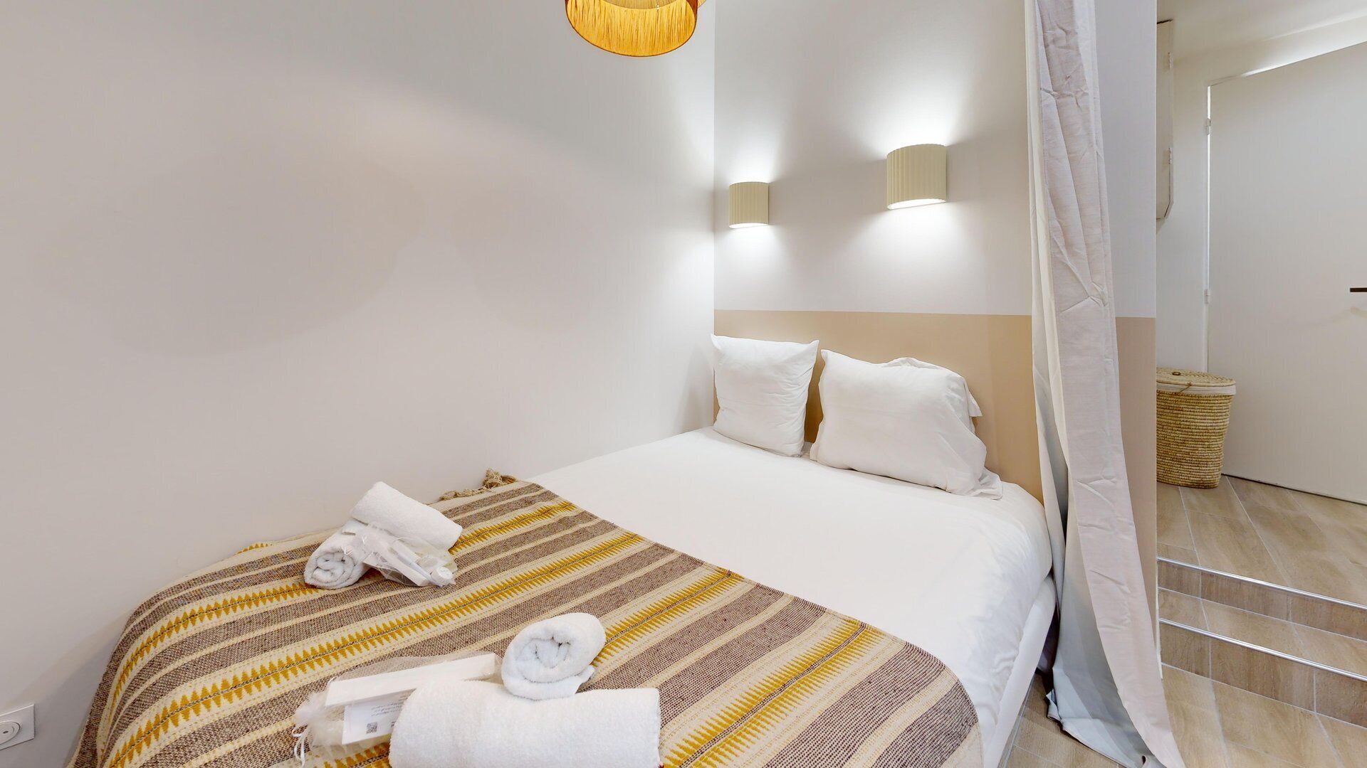 Een hotelkamer met een bed, twee wandlampen, een hanglamp, een rieten mand en een gordijn.