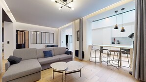 A modern living room with a grey sofa, a small round table, and a kitchen area with a dining table and bar stools. - TBS 151- Lovely suite near Place de la République (Paris)