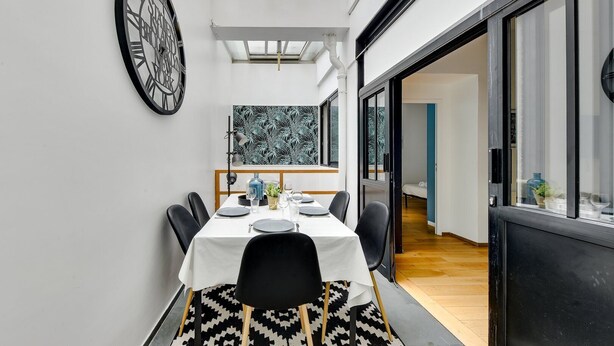 A modern dining area with a table set for four, a large clock on the wall, and a view of the adjacent room through a glass door. - TBS 6 - Beautiful apartment near Opéra Garnier (Paris)