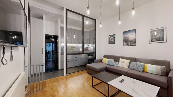 A modern living room with a sofa, a coffee table, and a TV mounted on the wall. There is a kitchen visible through a glass door, and the room has a minimalist design with a wooden floor. - TBS 6 - Beautiful apartment near Opéra Garnier (Paris)