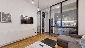A modern living room with a flat-screen TV, a sofa, a coffee table, and a kitchen visible through a glass door. - TBS 6 - Beautiful apartment near Opéra Garnier (Paris)