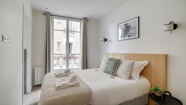 A modern hotel room with a large bed, a view of the city through a window, a wall-mounted light, and a framed picture.