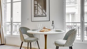 A dining area with a round table, two patterned chairs, a framed artwork on the wall, and a view of a building through large windows. - TheBlueStay 31 – Modern Apartment – Paris 13 (Paris)