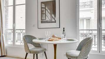 A dining area with a round table, two patterned chairs, a framed artwork on the wall, and a view of a building through large windows.