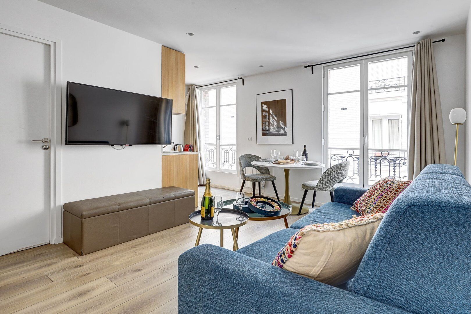 A modern living room with a blue sofa, a wooden bench, a flat-screen TV, a dining table with chairs, and a balcony with a railing.