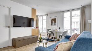 A modern living room with a blue sofa, a wooden bench, a flat-screen TV, a dining table with chairs, and a balcony with a railing. - TheBlueStay 31 – Modern Apartment – Paris 13 (Paris)