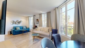 A modern living room with a blue sofa, a white sofa, and a glass coffee table. There are large windows with curtains, a picture on the wall, and a small table with a red object on it. - Bluestay 33 - Charming apartment in Paris (Paris)