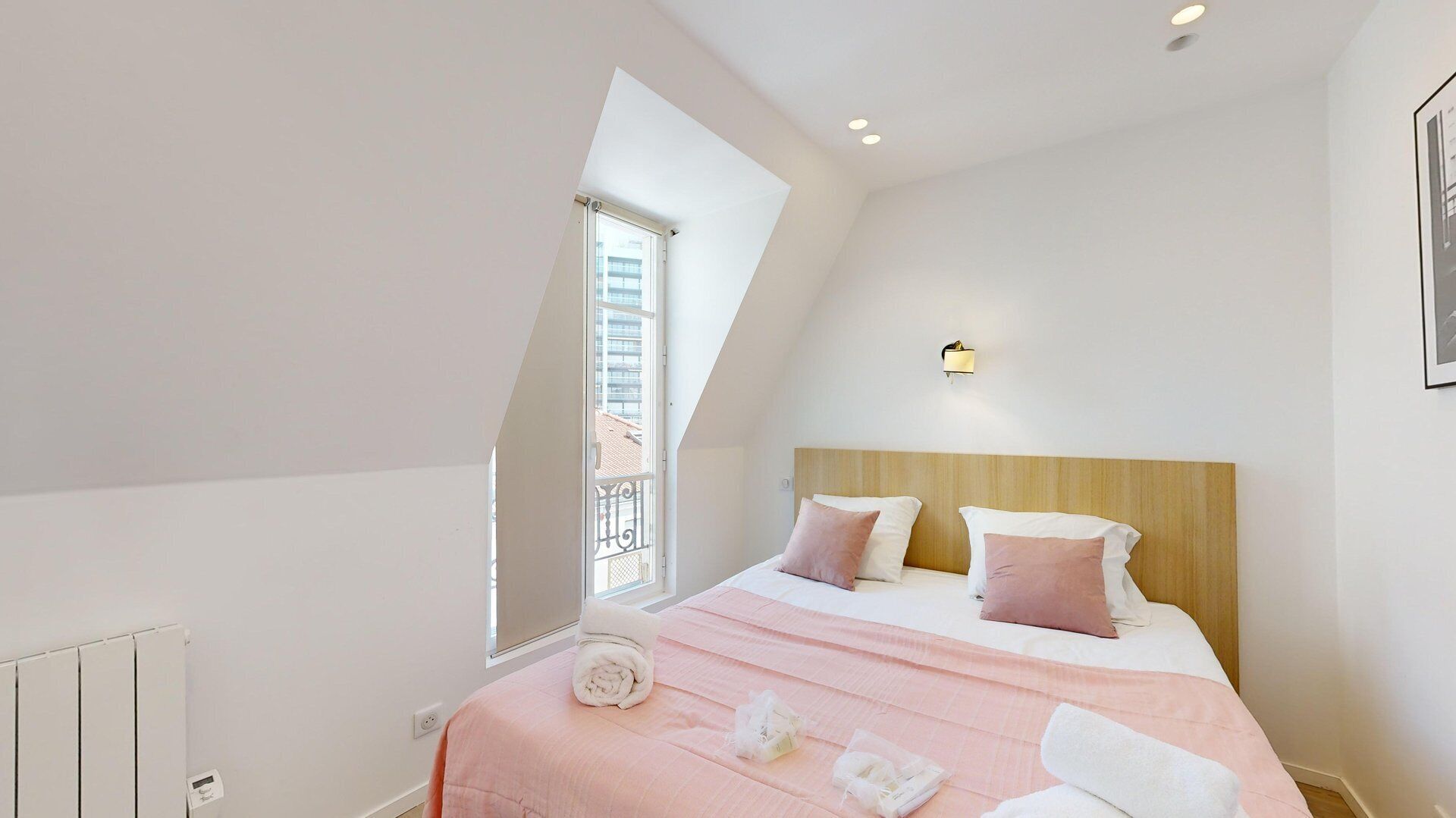 A modern bedroom with a sloped ceiling, a large window, a bed with pink bedding, and a wooden headboard.