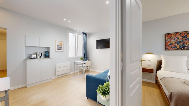 A modern, well-lit room with a bed, a desk, and a chair. There is a window with curtains, a flat-screen TV mounted on the wall, and a small table with a plant. - TBS 196 - Charming apartment in the Marais (Paris)