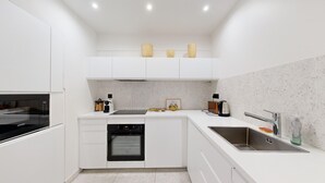 A modern kitchen with white cabinetry, a built-in oven, a stainless steel sink, and a countertop with a kettle and a framed picture. - TBS 43 - Sumptuous apartment in the heart of Paris (Paris)