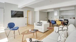 A modern living room with a white sofa, blue armchair, and a dining area with a table and chairs. A TV is mounted on the wall, and a kitchen is visible in the background. - TBS 43 - Sumptuous apartment in the heart of Paris (Paris)
