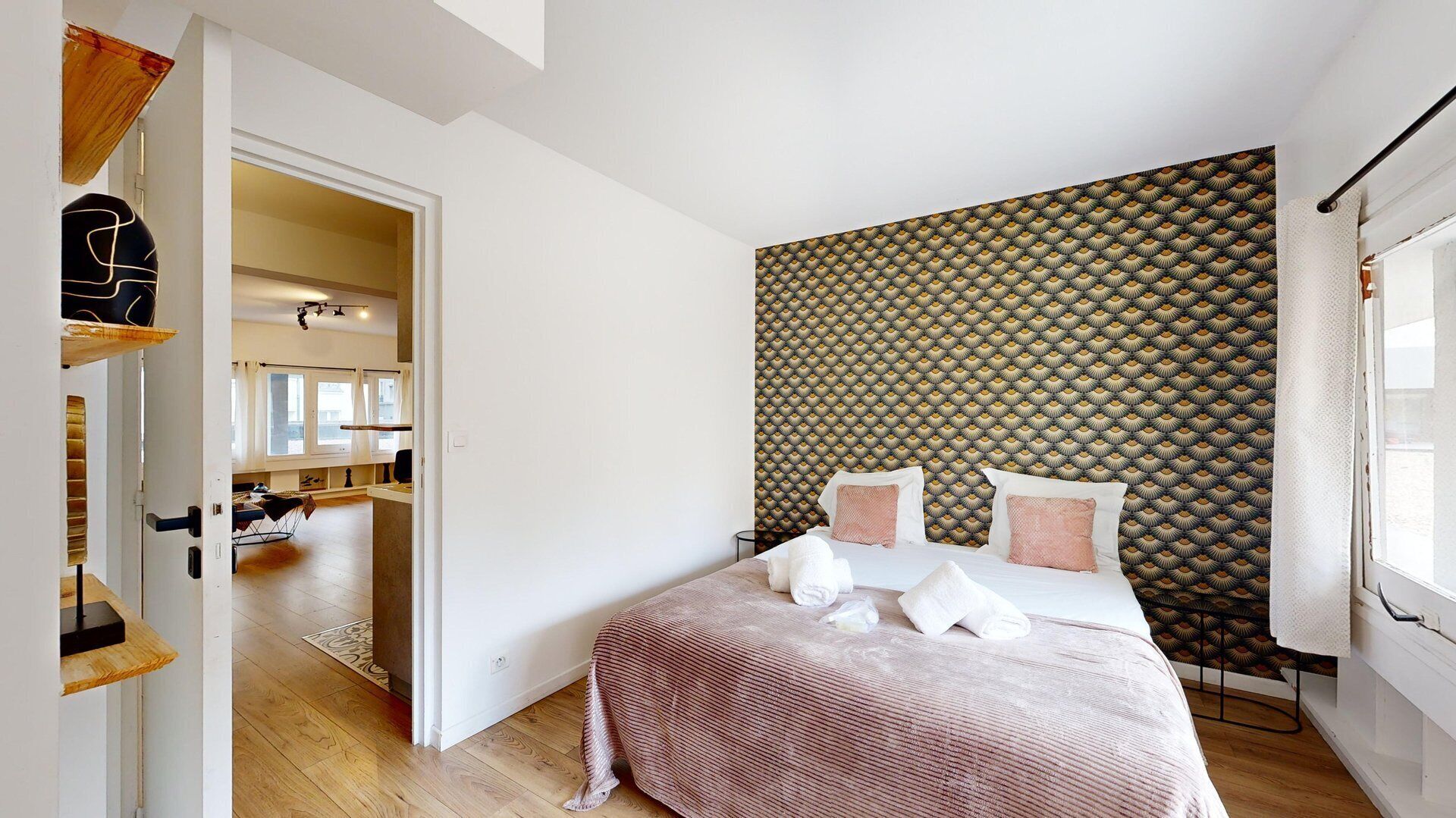 A bedroom with a patterned headboard, a bed with white linens and pink pillows, and a view of a hallway and another room through an open door.