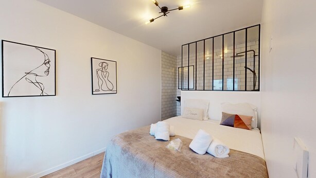 Een moderne slaapkamer met een groot bed, twee kunstwerken aan de muur, een plafondlamp en een badkamer met een glazen douchecabine.
