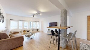Een ruime woonkamer met een groot raam, een bank, een salontafel en een eethoek met stoelen en een televisie.