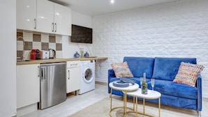 A compact living space with a kitchenette, a washing machine, a blue sofa with colorful cushions, and a small round table with a bottle and glasses.