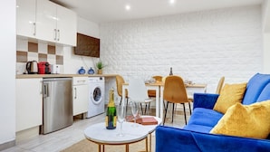 A modern kitchen with a washing machine, a dining area with a table and chairs, and a blue sofa with yellow cushions.