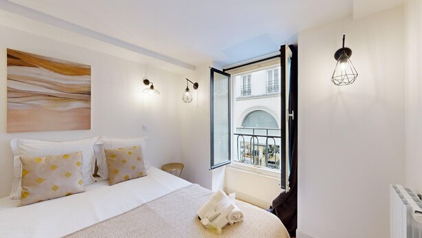 A bedroom with a bed, two pillows, a bedside table, a window with a view of buildings, and a wall-mounted light fixture.