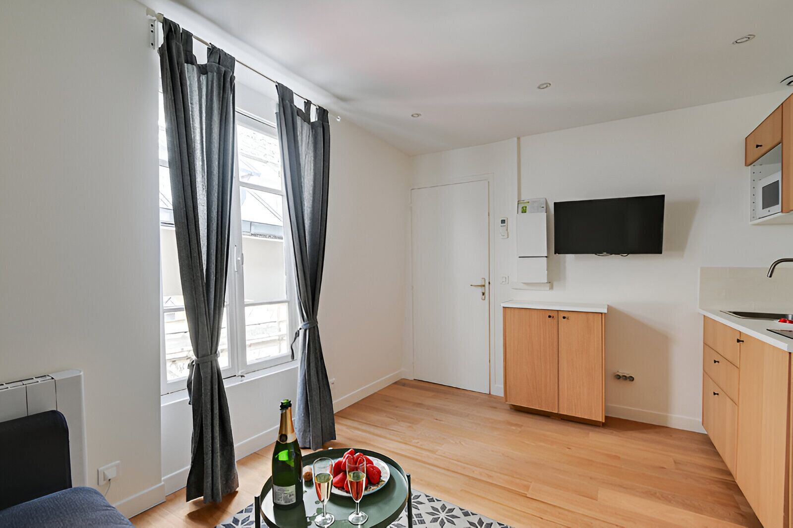 Een modern appartement met een houten vloer, een eethoek met tafel en stoelen, een keuken met een magnetron en een tv boven het aanrecht, en een raam met gordijnen.