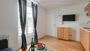 A modern apartment with a wooden floor, a dining area with a table and chairs, a kitchen with a microwave and a TV mounted above the counter, and a window with curtains. - TBS 16 - Elegant studio in Montorgueil district (Paris)