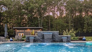 Una piscina con una cascada, rodeada por un muro de piedra y un edificio moderno al fondo.