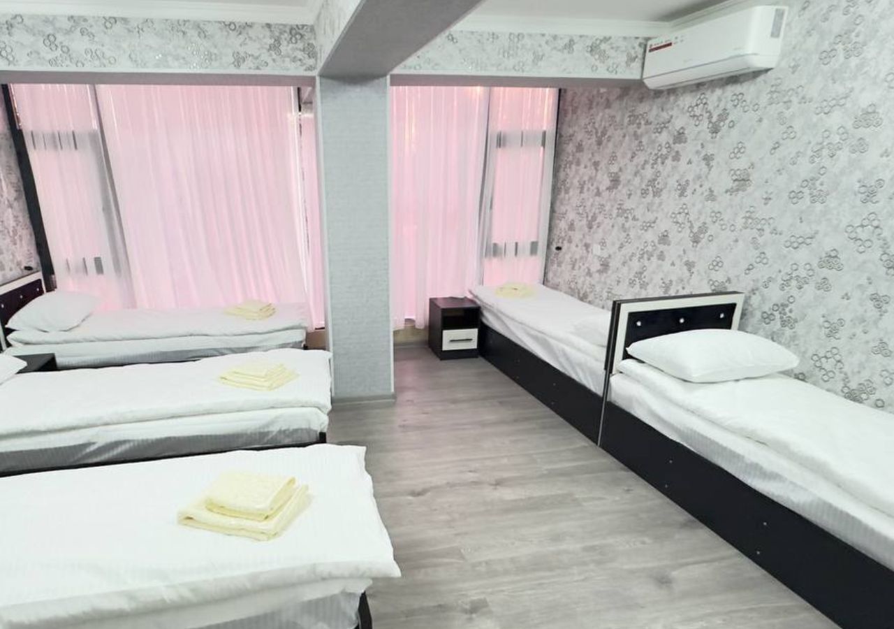 A hotel room with three single beds, a wooden floor, a small bedside table, and a window with pink curtains.