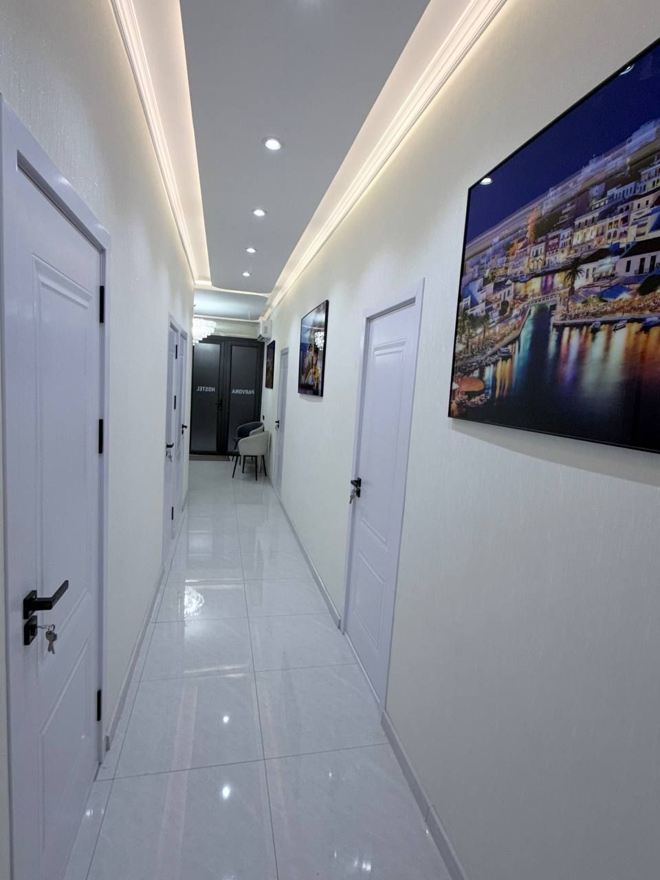 A hallway with white doors, a tiled floor, recessed lighting, and a large cityscape painting on the wall.