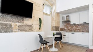 Een moderne keuken met witte kasten, een stenen muur, een flatscreen televisie en een klein eethoekje met twee stoelen en een tafel.