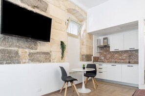 A modern kitchen with white cabinets, a stone wall, a flat-screen TV, and a small dining area with two chairs and a table.